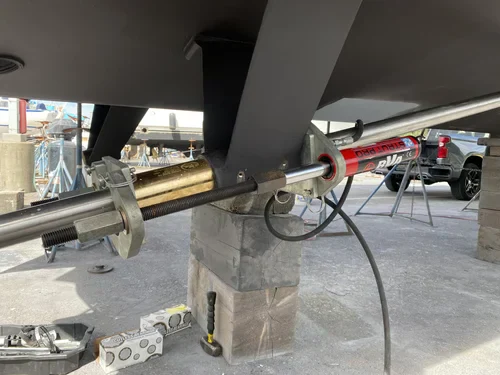 A hydraulic cylinder and other machinery are attached to the underside of a boat's hull, which is propped up on blocks