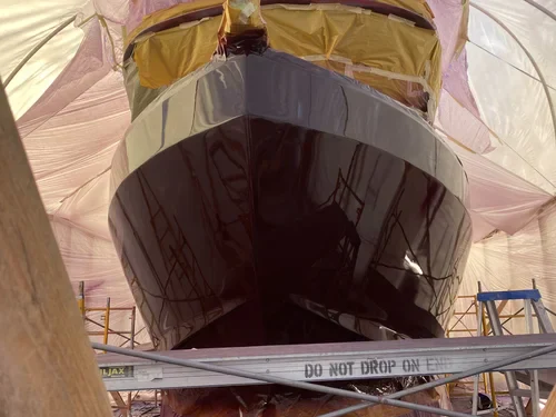 A close-up of a dark-hulled boat inside a temporary shelter with scaffolding, appearing to be in a finishing or painting process
