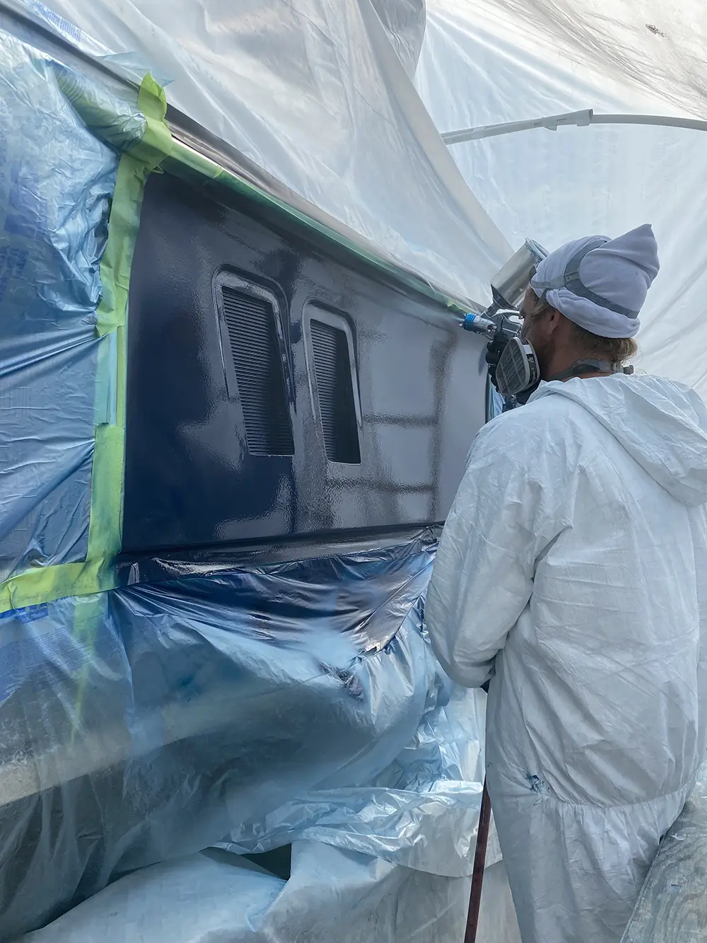 A person in protective gear spray paints a dark blue panel on a masked surface.