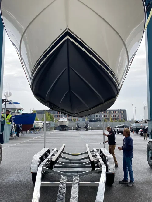 A white boat is being lowered from a travel lift onto a trailer at a boatyard. Two men are standing near the trailer, observing the process