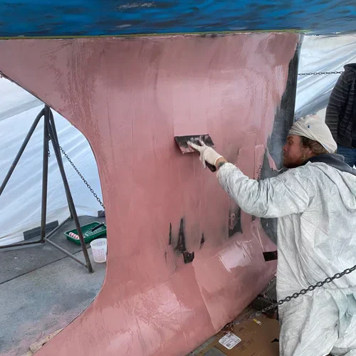 A person in a protective suit applies a pink substance to the hull of a boat with a trowel